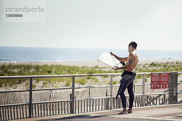 Mann sieht Surfbrett an  während er auf der Brücke gegen das Meer steht