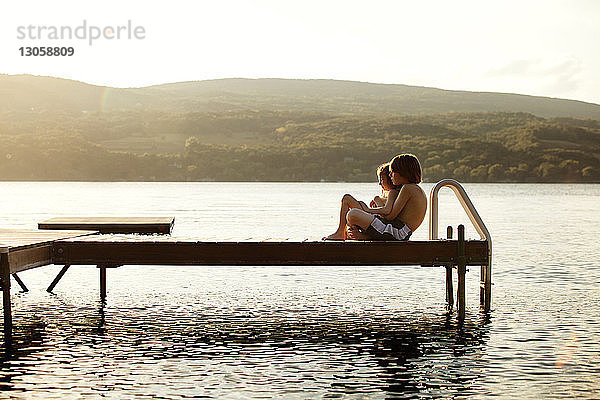 Seitenansicht der Geschwister  die an einem sonnigen Tag am Steg über dem See sitzen