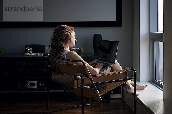 Frau benutzt Laptop  während sie zu Hause auf einem Stuhl sitzt