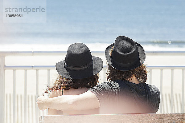Rückansicht eines Paares  das Hüte trägt  während es am Strand auf einer Bank sitzt