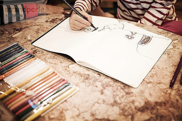 Beschnittenes Bild eines Jungen  der zu Hause bei Tisch auf ein Buch zeichnet
