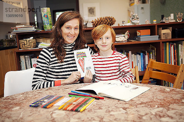 Porträt von Mutter und Sohn  die eine Karte halten  während sie auf einem Stuhl am Tisch sitzen