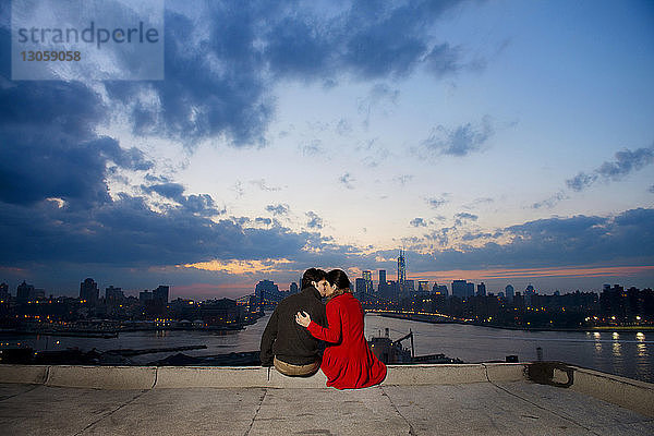 Rückansicht eines jungen Paares  das sich in der Abenddämmerung gegen Fluss und Stadtbild küsst