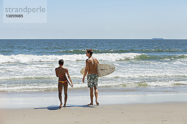 Rückansicht eines Surfbrett tragenden Paares am Strand