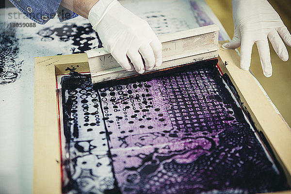 Ausgeschnittene Hände einer Handwerkerin  die im Siebdruckverfahren das Design in der Werkstatt auf Stoff druckt