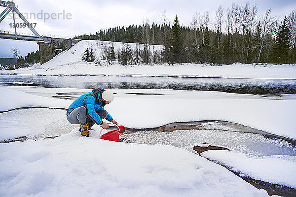 Mann füllt Eimer  während er am schneebedeckten Flussufer kauert