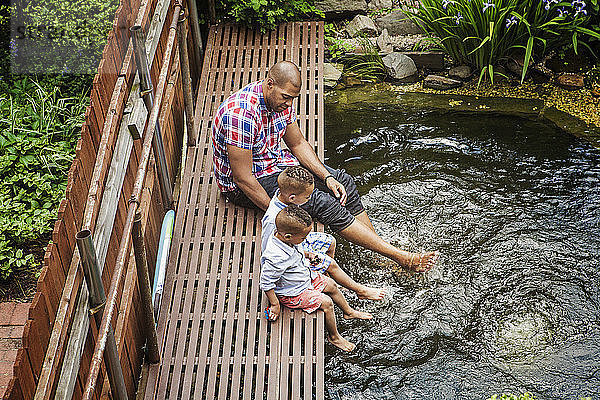 Hochwinkelaufnahme eines Vaters mit Söhnen  die auf einem Fußweg über einem Teich im Hinterhof sitzen