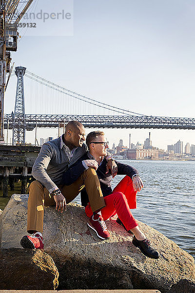 Schwule Männer schauen auf East River  während sie auf Felsen sitzend gegen die Williamsburg-Brücke blicken