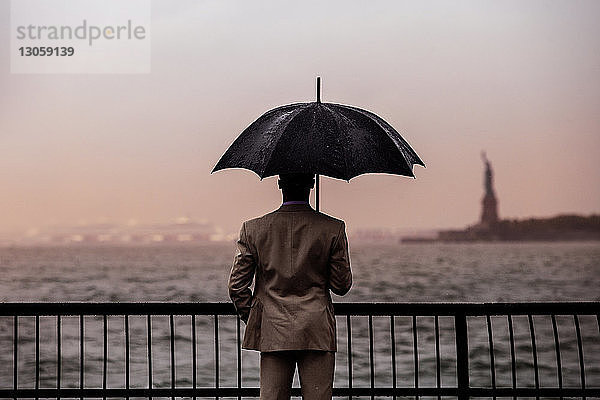 Rückansicht eines Mannes mit Regenschirm  der an der Reling gegen das Meer steht