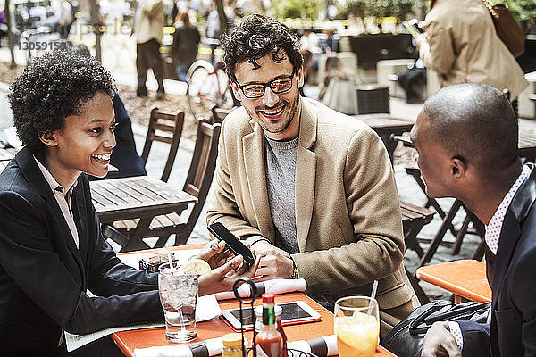 Glückliche Geschäftsfrau zeigt Geschäftsleuten ihr Smartphone  während sie im Straßencafé sitzt