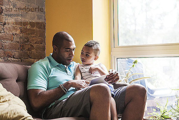 Mann benutzt Tablet-Computer  während er mit seinem Sohn zu Hause auf dem Sofa sitzt