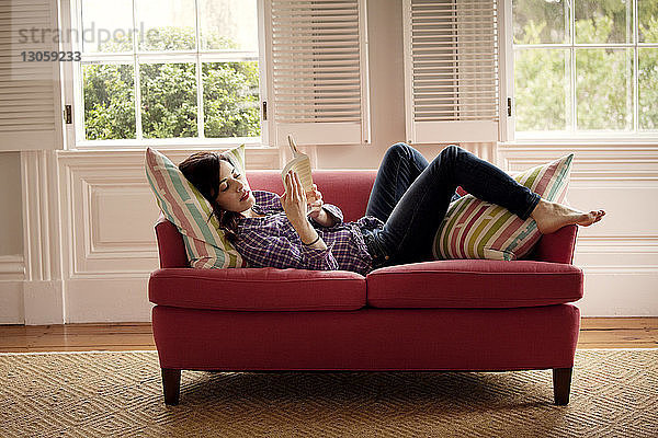 Frau liest Buch  während sie zu Hause auf dem Sofa liegt