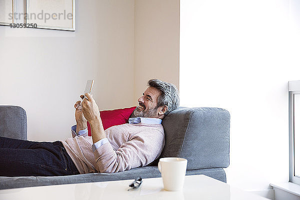 Lächelnder Geschäftsmann schaut auf Tablet-Computer  während er auf dem Sofa liegt