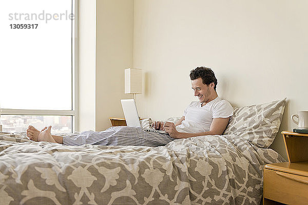Seitenansicht eines glücklichen Mannes  der seinen Laptop zu Hause auf dem Bett benutzt