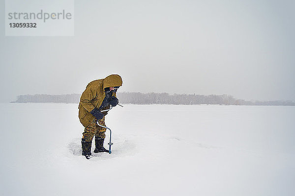 Älterer Mann bohrt in zugefrorenem See gegen klaren Himmel