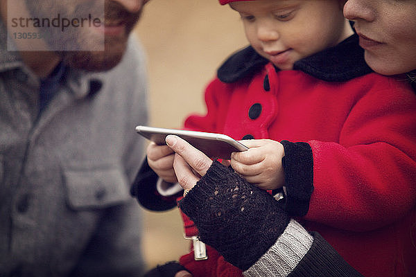 Kleines Mädchen mit Eltern  die im Park auf ein Mobiltelefon schauen