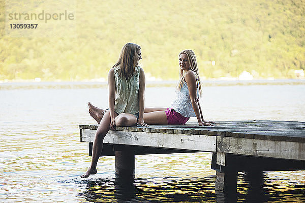 Lächelnde Freunde unterhalten sich  während sie auf dem Steg über dem See sitzen