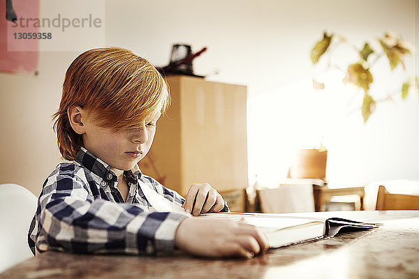 Junge liest Buch  während er zu Hause am Tisch sitzt