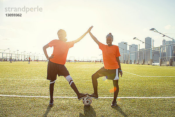 Porträt von glücklichen Sportlerinnen  die bei strahlendem Sonnenschein mit dem Fussball auf dem Spielfeld Abklatschen