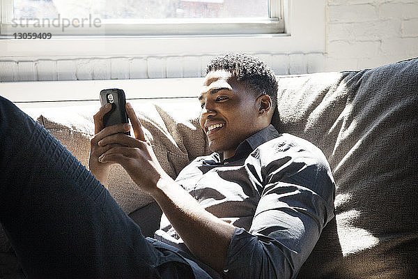 Lächelnder Mann benutzt Smartphone  während er zu Hause auf dem Sofa liegt