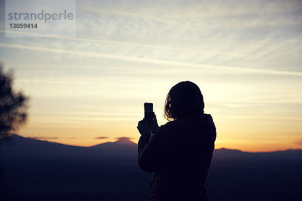 Silhouettenfrau fotografiert mit Smartphone gegen den Himmel bei Sonnenuntergang