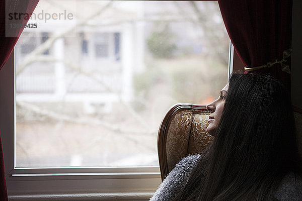 Nachdenkliche Frau schaut durch das Fenster  während sie zu Hause auf einem Stuhl sitzt