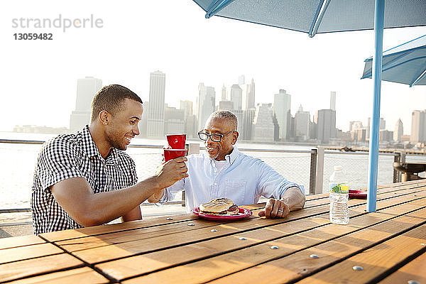 Glückliche Vater und Sohn stoßen auf Getränke an  während sie am East River in der Stadt am Tisch sitzen