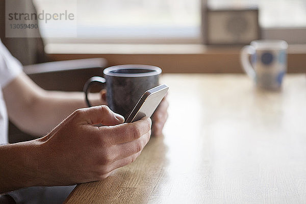 Ausgeschnittenes Bild eines Mannes  der ein Mobiltelefon benutzt  während er im Café eine Kaffeetasse hält