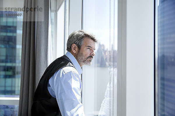 Nachdenklicher Geschäftsmann schaut durch Fenster  während er im Hotelzimmer steht