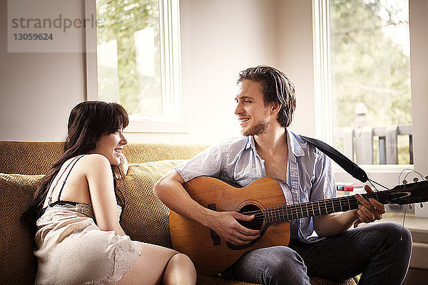 Mann spielt Gitarre  während er zu Hause mit seiner Freundin auf dem Sofa sitzt