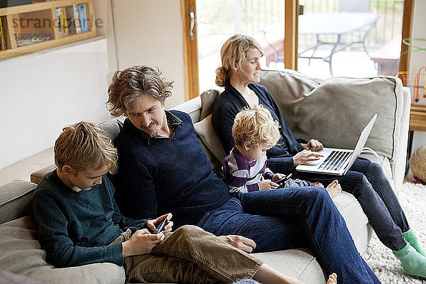 Familie benutzt Laptop und Mobiltelefon  während sie zu Hause auf dem Sofa sitzt