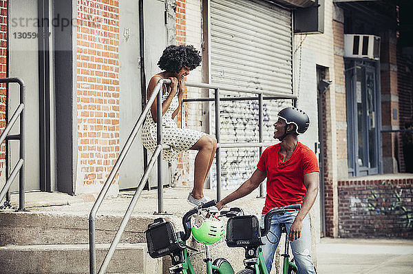 Mann mit Fahrrad schaut Frau an  die auf einem Geländer sitzt