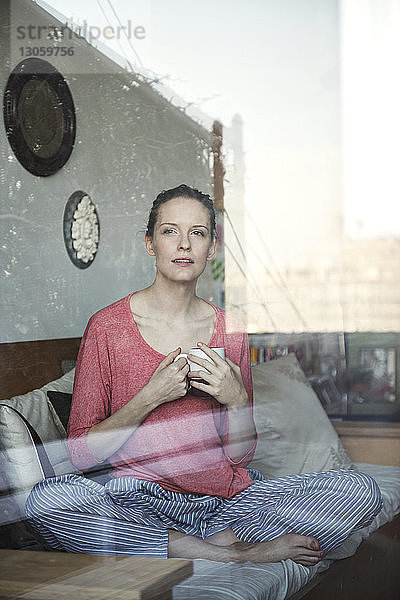 Frau hält Kaffeetasse und sitzt auf Sofa durch Glasfenster gesehen