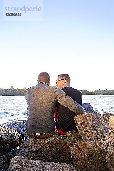 Rückansicht eines glücklichen Mannes  der mit seinem Freund auf Felsen am East River vor klarem Himmel Arm in Arm mit seinem Freund sitzt