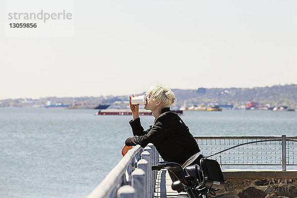 Frau trinkt Kaffee  während sie sich am Beobachtungspunkt am Fluss an ein Geländer lehnt