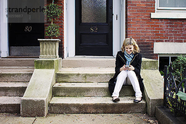 Frau benutzt ein Smartphone  während sie auf einer Treppe vor dem Haus sitzt