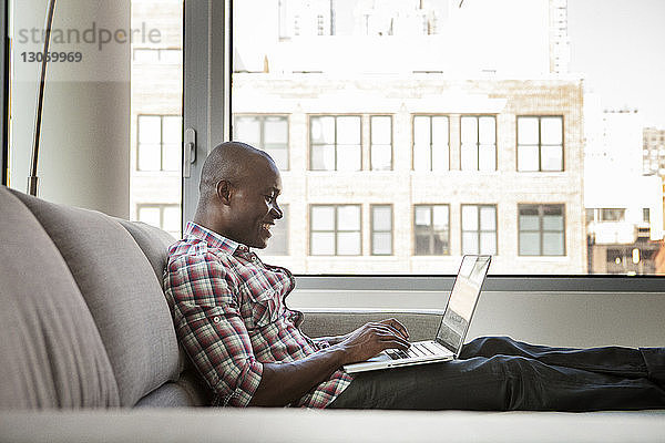 Seitenansicht eines glücklichen Mannes  der einen Laptop-Computer benutzt  während er zu Hause auf dem Sofa sitzt