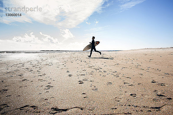 Seitenansicht eines Mannes mit Surfbrett  der auf Sand am Strand vor bewölktem Himmel läuft