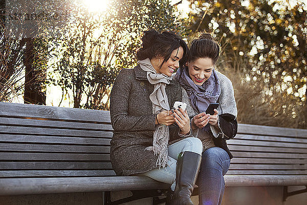 Lächelnde Frauen  die Smartphones benutzen  während sie auf einer Bank sitzen