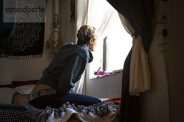 Seitenansicht einer Frau  die durch das Fenster schaut  während sie zu Hause auf dem Bett sitzt