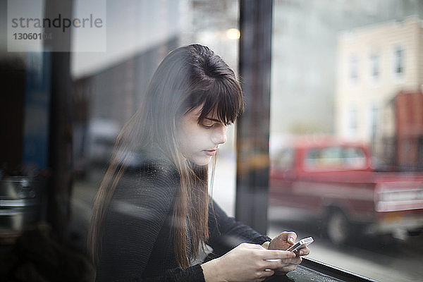 Nahaufnahme einer jungen Frau  die ein Smartphone benutzt  durch ein Fenster gesehen