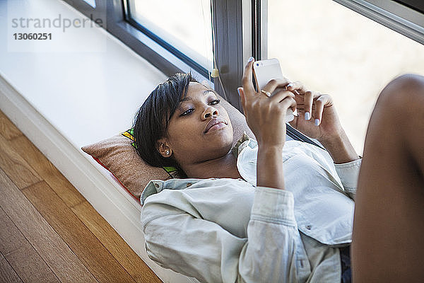 Frau benutzt Mobiltelefon  während sie zu Hause am Fenster liegt