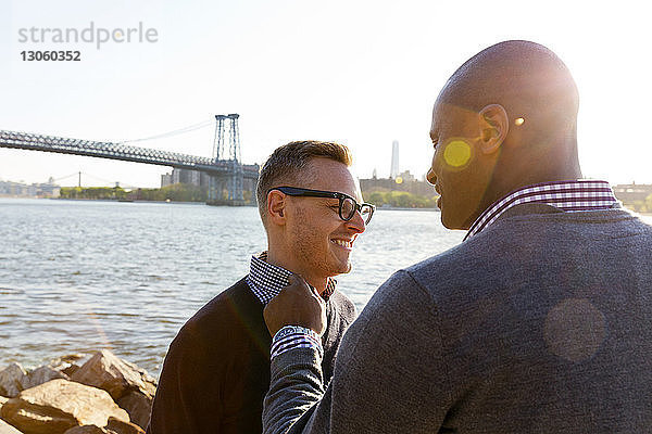 Lächelnder Mann berührt den Kragen seines Freundes  während er am East River steht