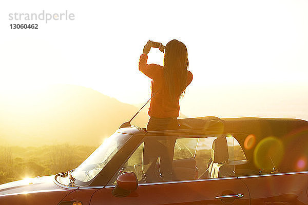 Rückansicht einer Frau  die fotografiert  während sie am Autoschiebedach steht