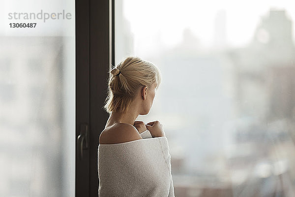 Seitenansicht einer in ein Handtuch gehüllten Frau  die zu Hause am Fenster steht
