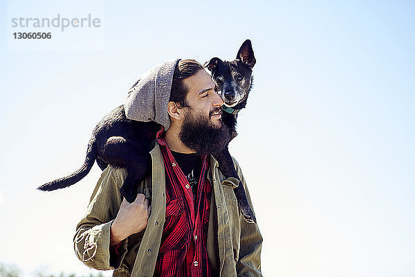 Nachdenklicher Mann trägt Hund auf den Schultern  während er sich gegen den klaren Himmel stellt