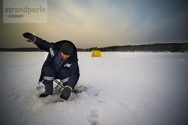 Ausgewachsene männliche Eisfischerei auf gefrorenem See gegen den Himmel bei Sonnenuntergang