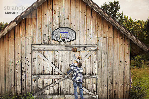 Rückansicht eines Vaters  der einen Sohn trägt  während er vor der Scheune Basketball spielt