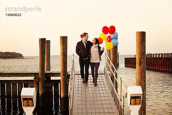 Frau hält Heliumballons  während sie mit dem Mann bei Sonnenuntergang am Pier spazieren geht