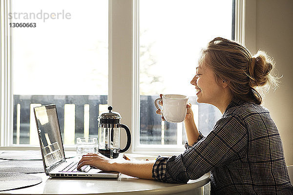 Glückliche Frau benutzt Laptop-Computer  während sie Kaffee am Tisch genießt